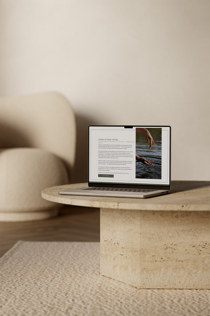 Laptop displaying an article with a swimmer photo sits on a round stone coffee table in a warm, minimalist living room beside a cream chair.