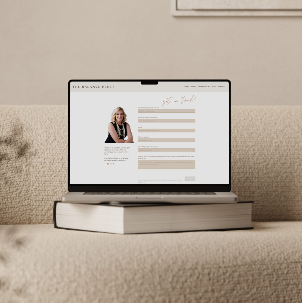 A laptop displaying a polished personal website rests on a thick book on a beige sofa; the neutral tones and tidy scene feel calm and professional.