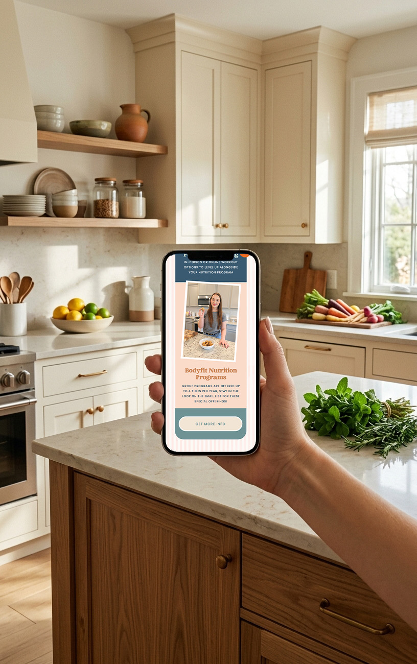 Hand holds a smartphone displaying a nutrition program in a bright farmhouse kitchen, with fresh vegetables, herbs, jars, and wooden cabinets.