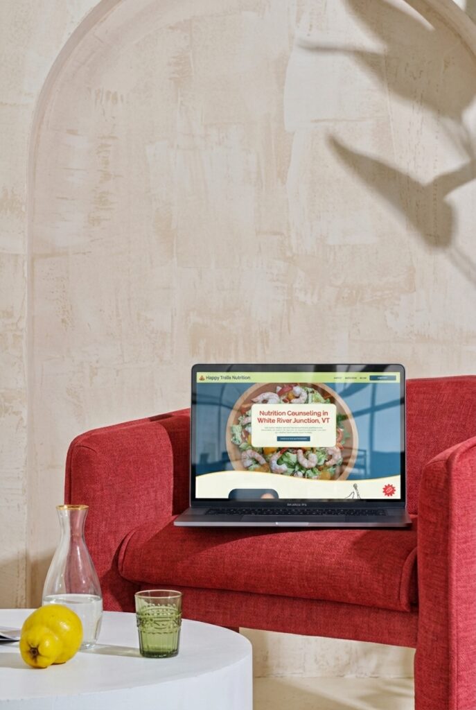 Laptop displaying a nutrition counseling website on a red sofa, with lemon water and a glass on a white table; bright, calm interior.