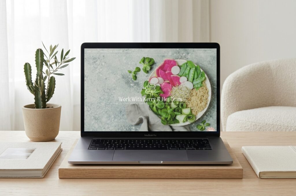Laptop on a wooden desk displays a colorful salad bowl with “Work With Kerry & Her Team”; a potted cactus and notebooks create a calm office scene.