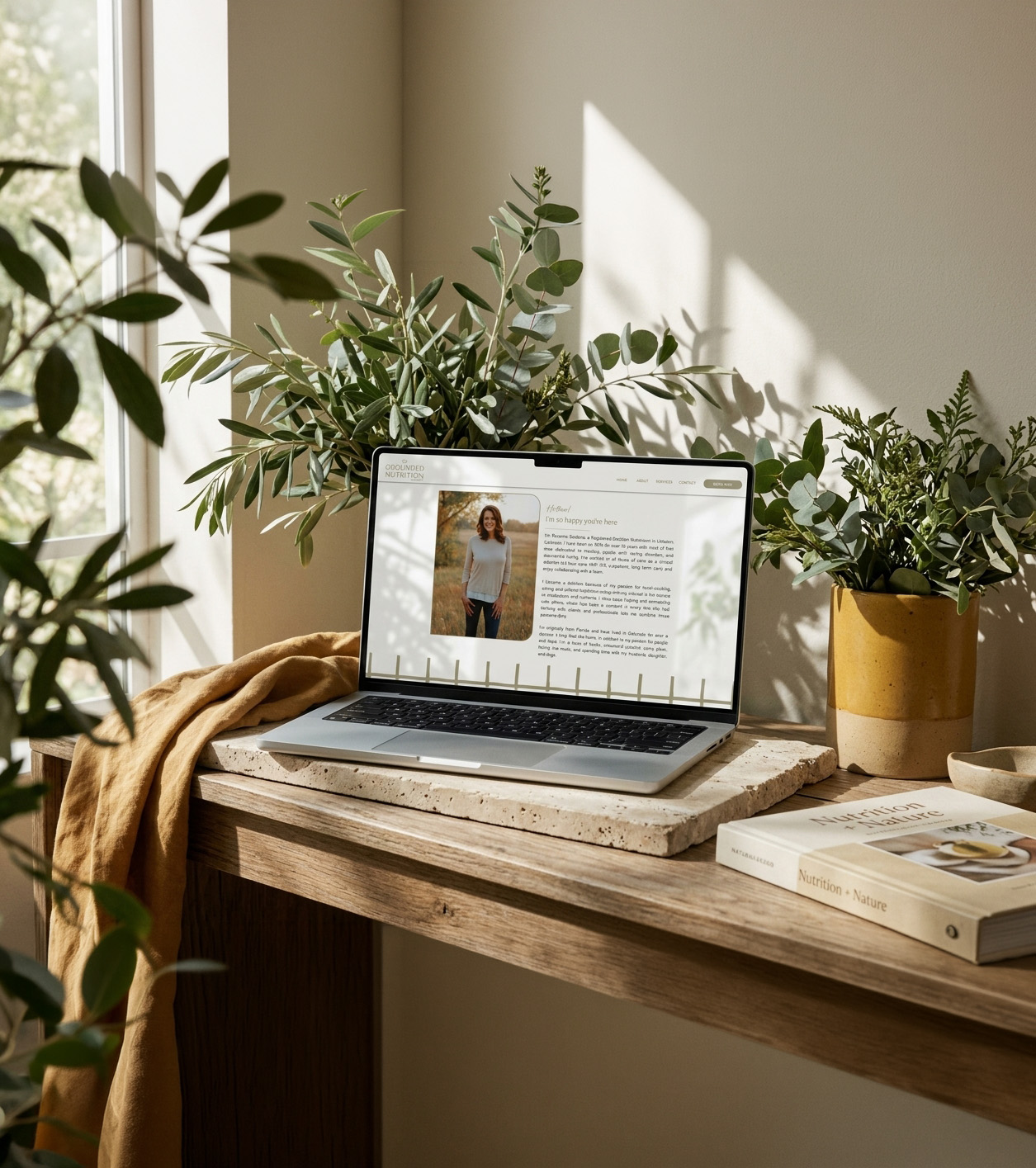 Sunlit home office with a laptop displaying a profile webpage, surrounded by leafy plants, books, and a cozy blanket on a wooden desk.