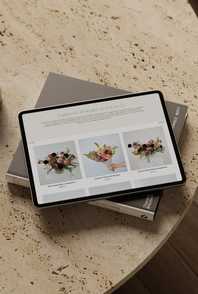 Tablet displaying a floral design website with three bouquet arrangements, resting on books atop a warm-toned stone table; elegant, calm setting.