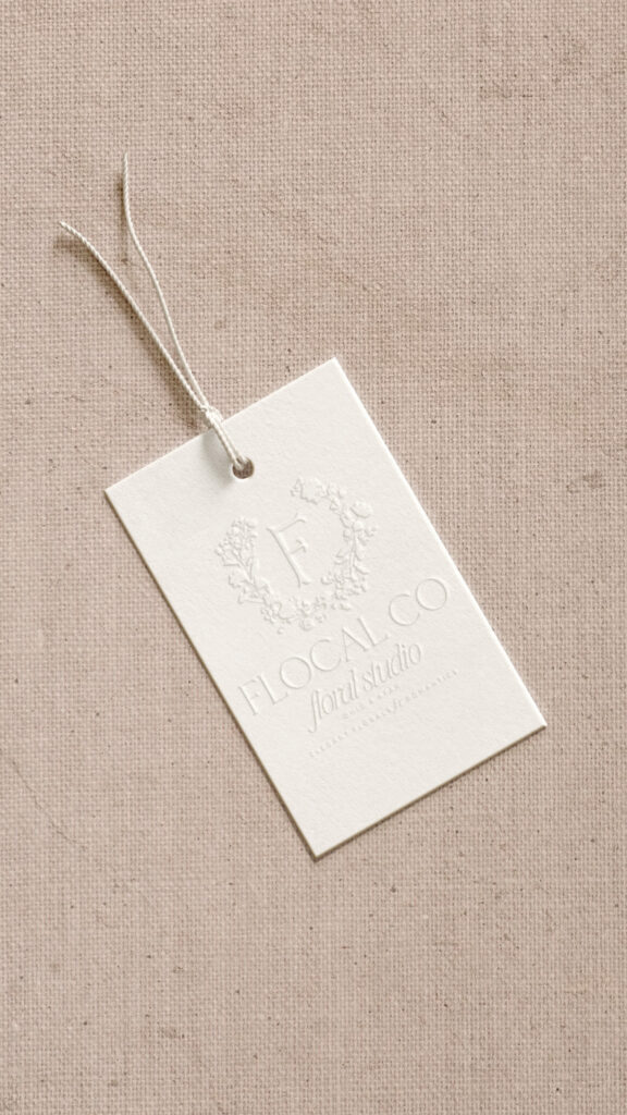 White clothing tag reading “Flocal Co.” tied with string, rests on beige linen fabric; embossed floral details create a soft, minimalist mood.