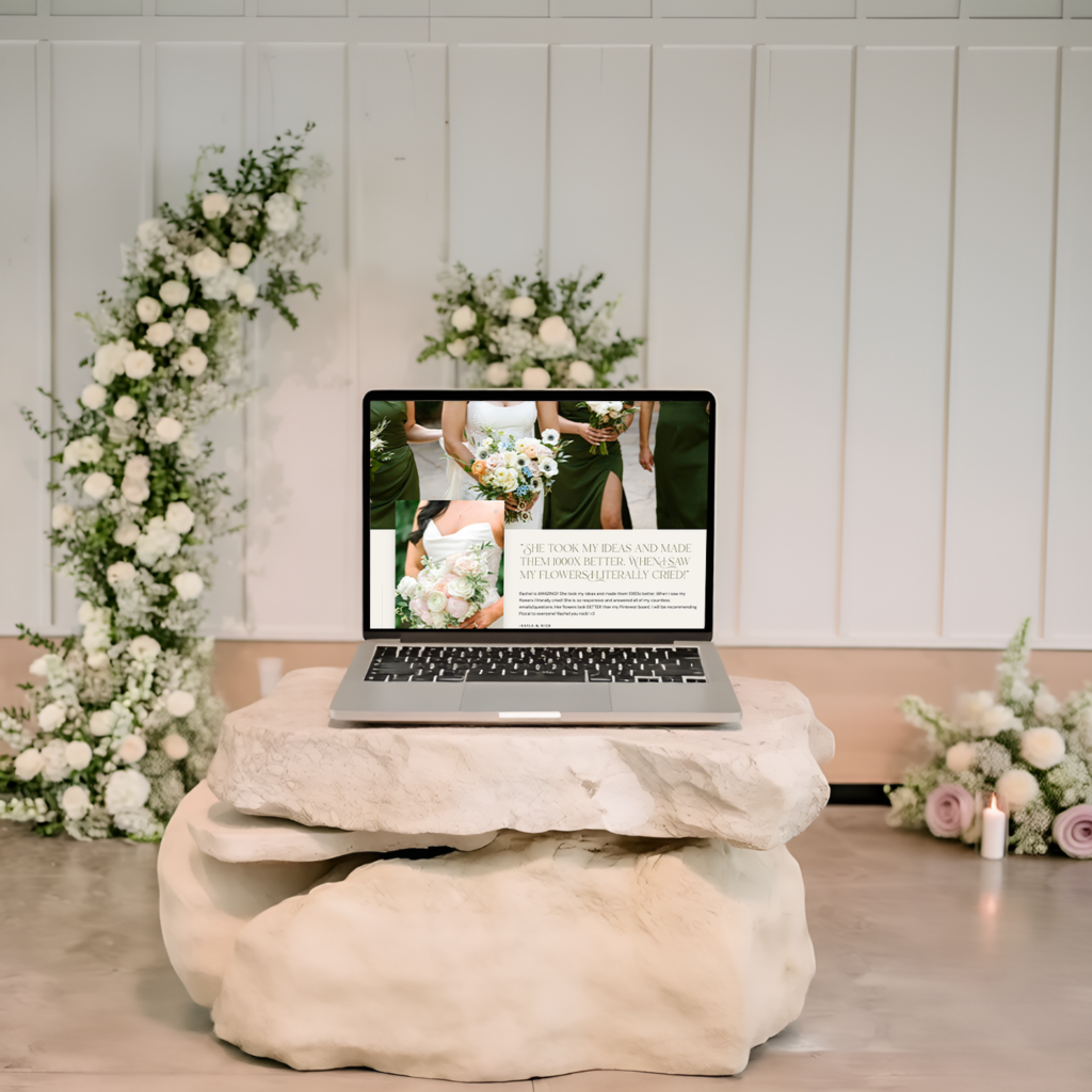 Laptop displaying a romantic wedding website on a stone pedestal, surrounded by white floral arrangements against a paneled backdrop; elegant, serene mood.