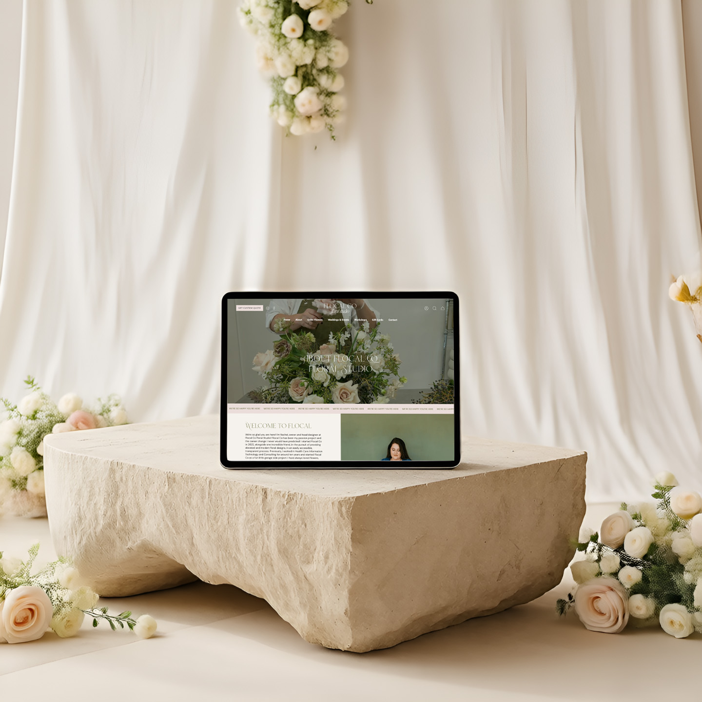 Tablet displaying a floral wedding website rests on a rough stone pedestal, surrounded by white roses and draped fabric; serene, elegant scene.