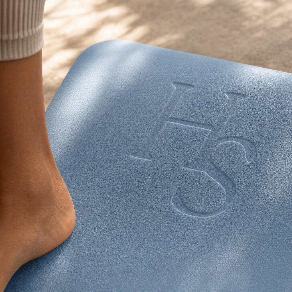 Close-up of a bare foot beside a blue textured mat embossed with “HS,” softly lit by sunlight, creating a calm, wellness-focused mood.