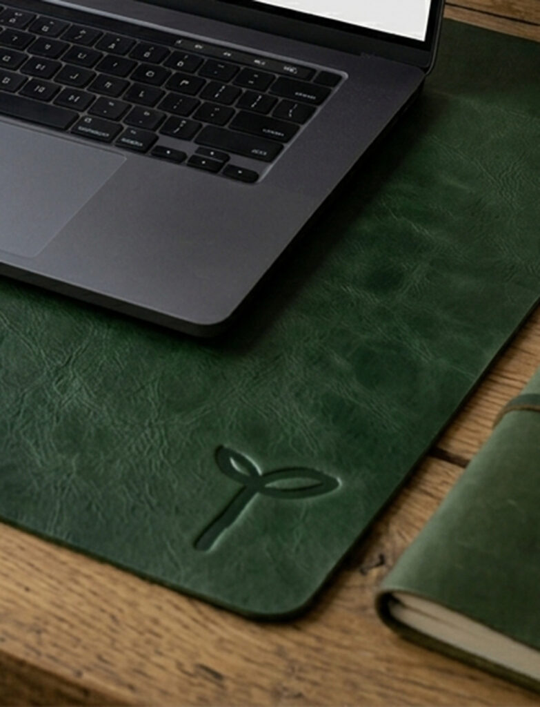 A silver laptop rests on a textured dark green leather desk mat embossed with a sprout, beside a matching notebook on a wooden desk; calm, refined workspace.