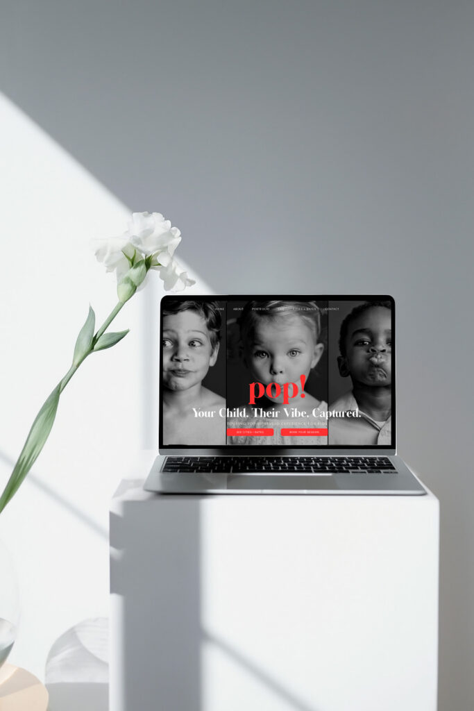 Minimalist scene with laptop on a white pedestal displaying black-and-white portraits of three children, red “pop!” text, flower and shadows.