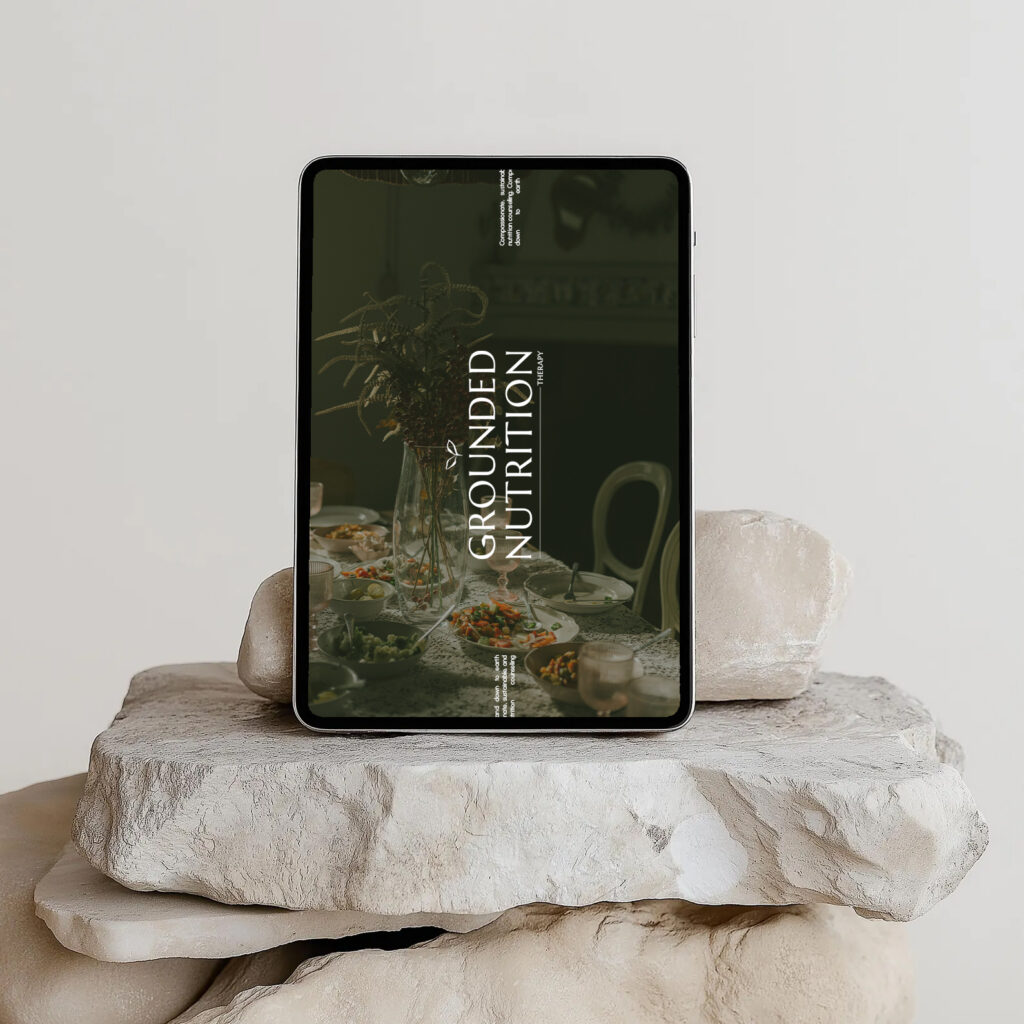Tablet displaying “Grounded Nutrition” rests on stacked stone slabs, with a dim rustic dining scene visible on its screen; calm, earthy mood.