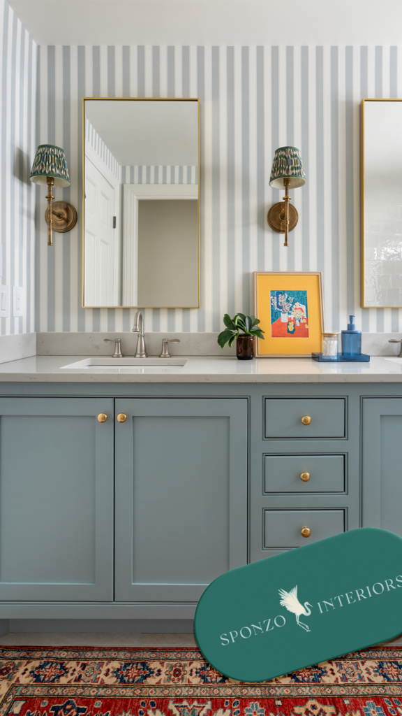 Elegant bathroom with blue-gray vanity, striped wallpaper, twin mirrors, brass sconces, colorful artwork, plants, and a patterned rug.