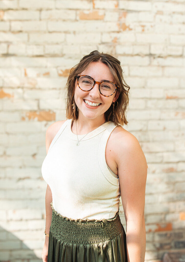 Smiling woman with round glasses and shoulder-length brown hair poses in a white sleeveless top and green skirt before a rustic brick wall.