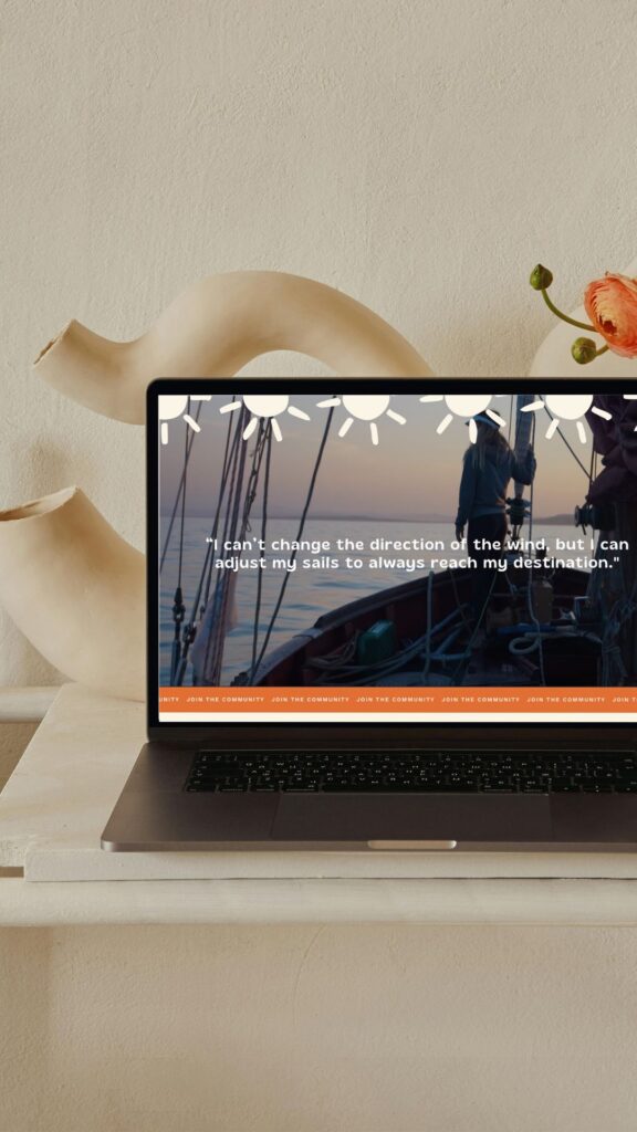 A laptop on a cream desk displays a sunlit forest scene with a person and motivational text, beside a sculptural vase and orange flower.
