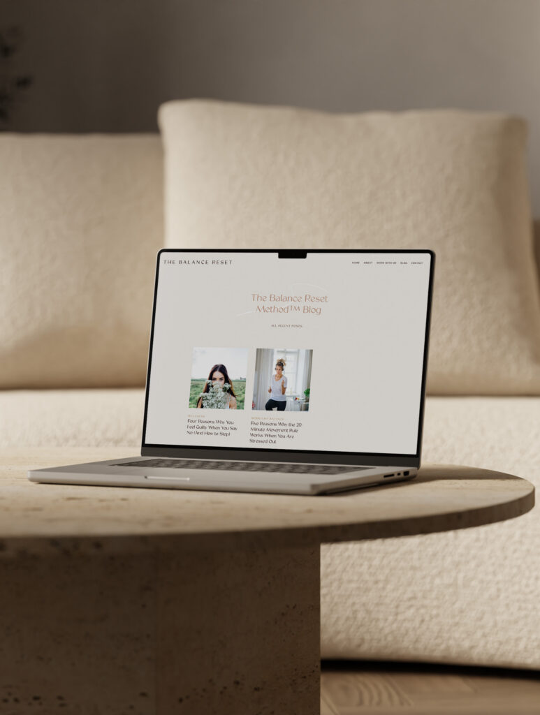 A laptop displays a minimalist fashion website on a round stone table before a cream sofa, creating a calm, warm, elegant living-room scene.