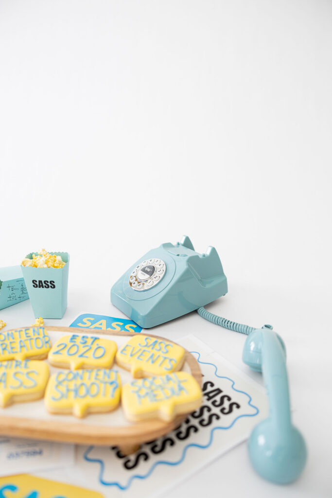 Aqua vintage rotary phone and handset sit beside yellow lettered cookies and event papers, with a tiny popcorn cup against a white backdrop; playful retro mood.