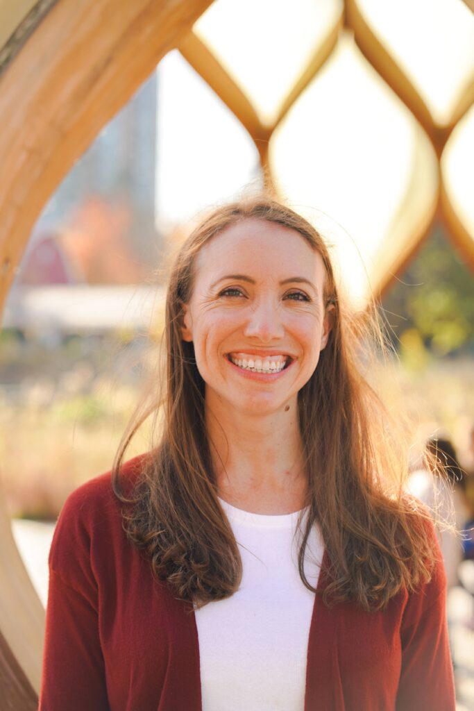 Portrait of a smiling woman in a red cardigan and white shirt, standing in warm sunlight beneath a geometric wooden canopy.