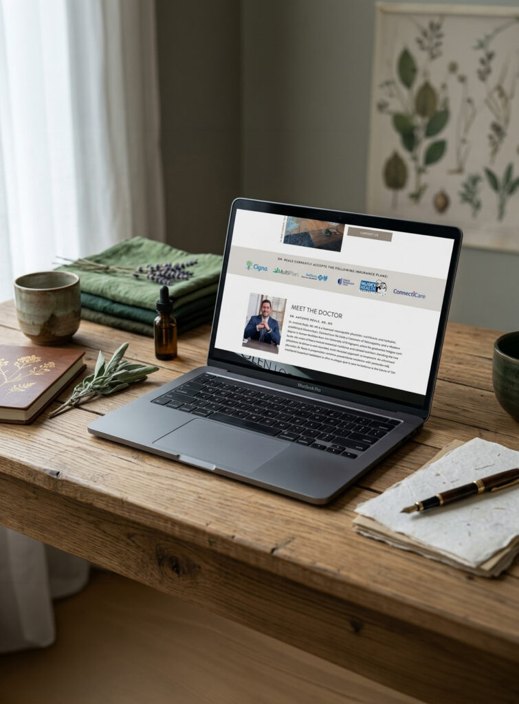 A laptop displays a wellness or medical website on a rustic wooden desk, surrounded by a mug, books, herbs, and a notebook, creating a calm, natural workspace.