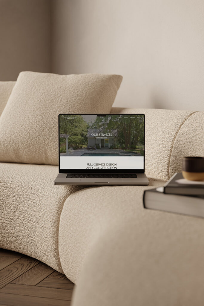 Laptop displaying a full-service interior design website rests on a cream sofa beside coffee and books, creating a warm, calm minimalist living room.