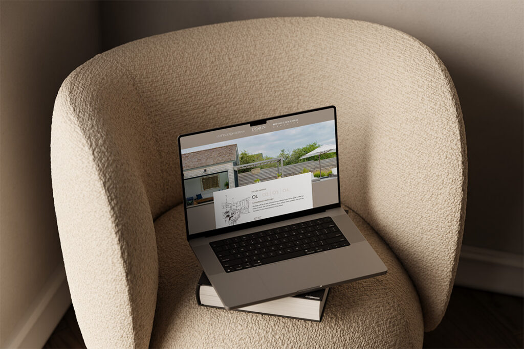 A laptop displaying a home website rests on a beige boucle armchair with papers beneath, creating a warm, quiet workspace scene in soft light.