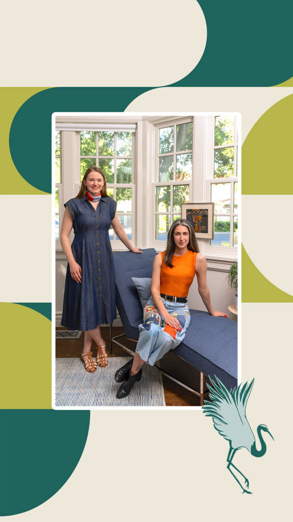 Two interior designers pose in a sunlit living room with large windows: one stands in a blue dress, the other sits in orange; a heron decorates the frame.
