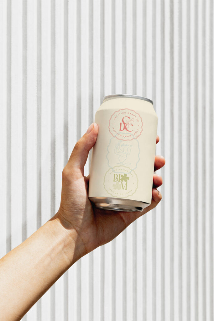 Hand holding a minimalist cream beverage can with pink and green floral lettering against a bright vertical striped background, calm and fresh.