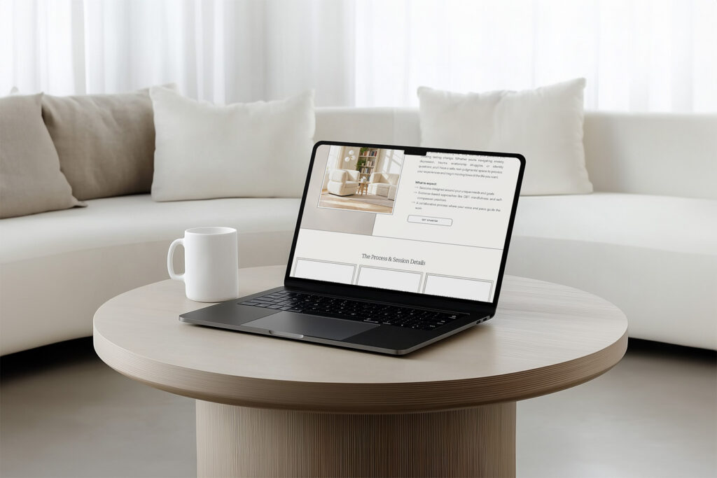 Laptop displaying a webpage on a round coffee table, beside a white mug, in a bright minimalist living room with a sofa; calm, modern mood.