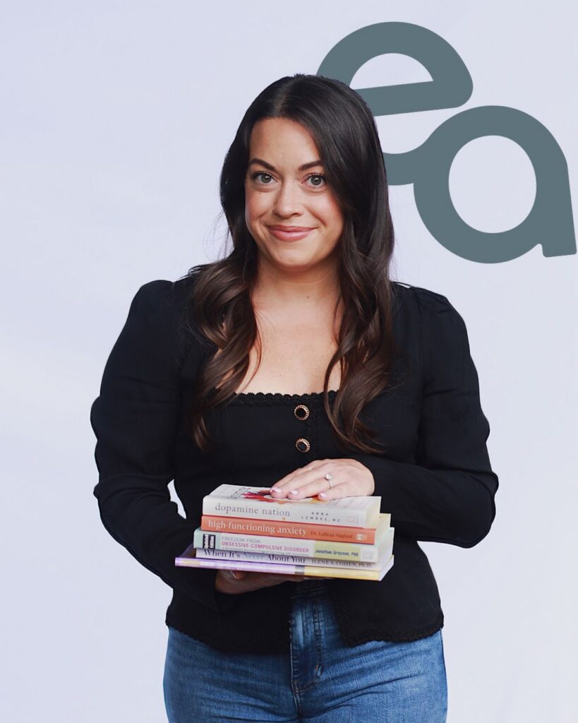 Smiling woman in black top and jeans holds a stack of books, posed before a gray abstract logo on a pale background; warm, confident tone.