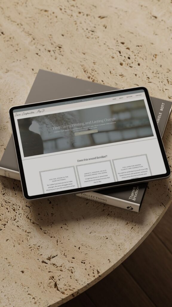 Tablet displaying a muted website rests on stacked design books atop a round stone table, creating a warm, sophisticated workspace scene.