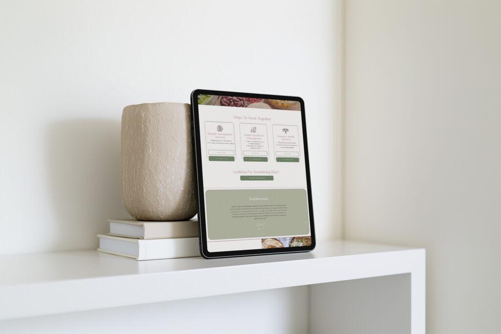 Tablet displaying a webpage rests against a beige ceramic vase and stacked books on a white shelf in a bright, minimalist room.