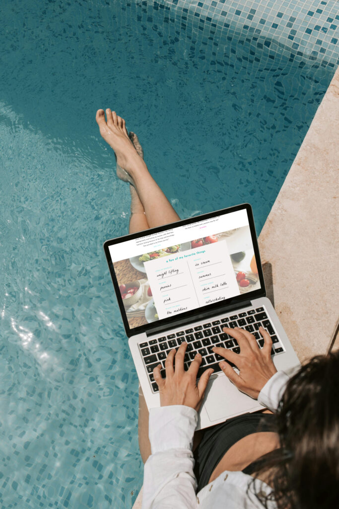 Person relaxes poolside with feet in turquoise water, typing on a laptop showing a webpage, blending work with a calm summer setting.