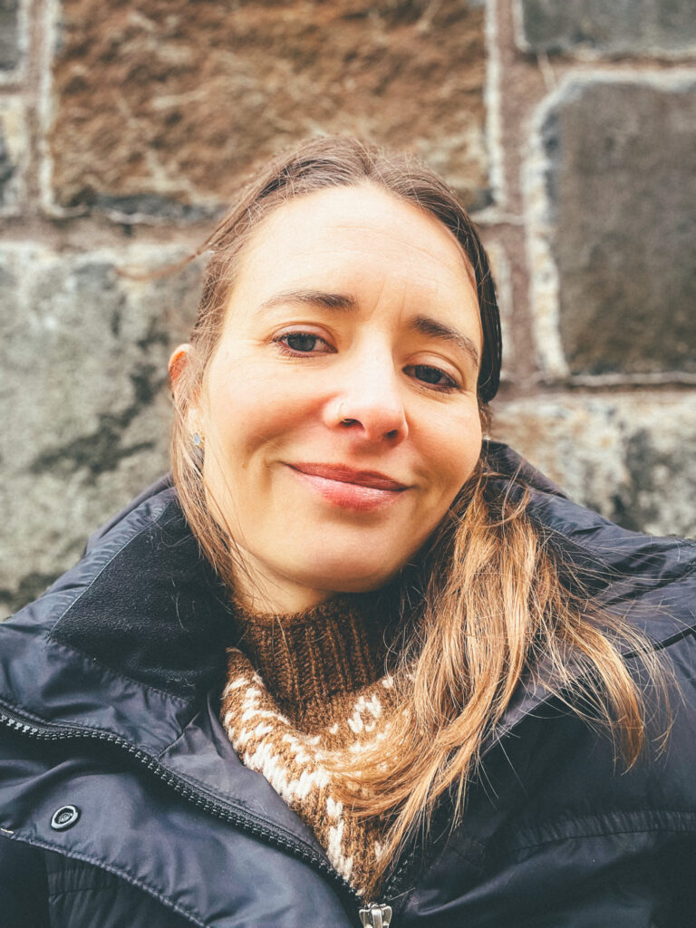 A woman in a black winter coat and brown patterned scarf smiles gently for a close-up outdoor portrait, with a weathered stone wall behind her.