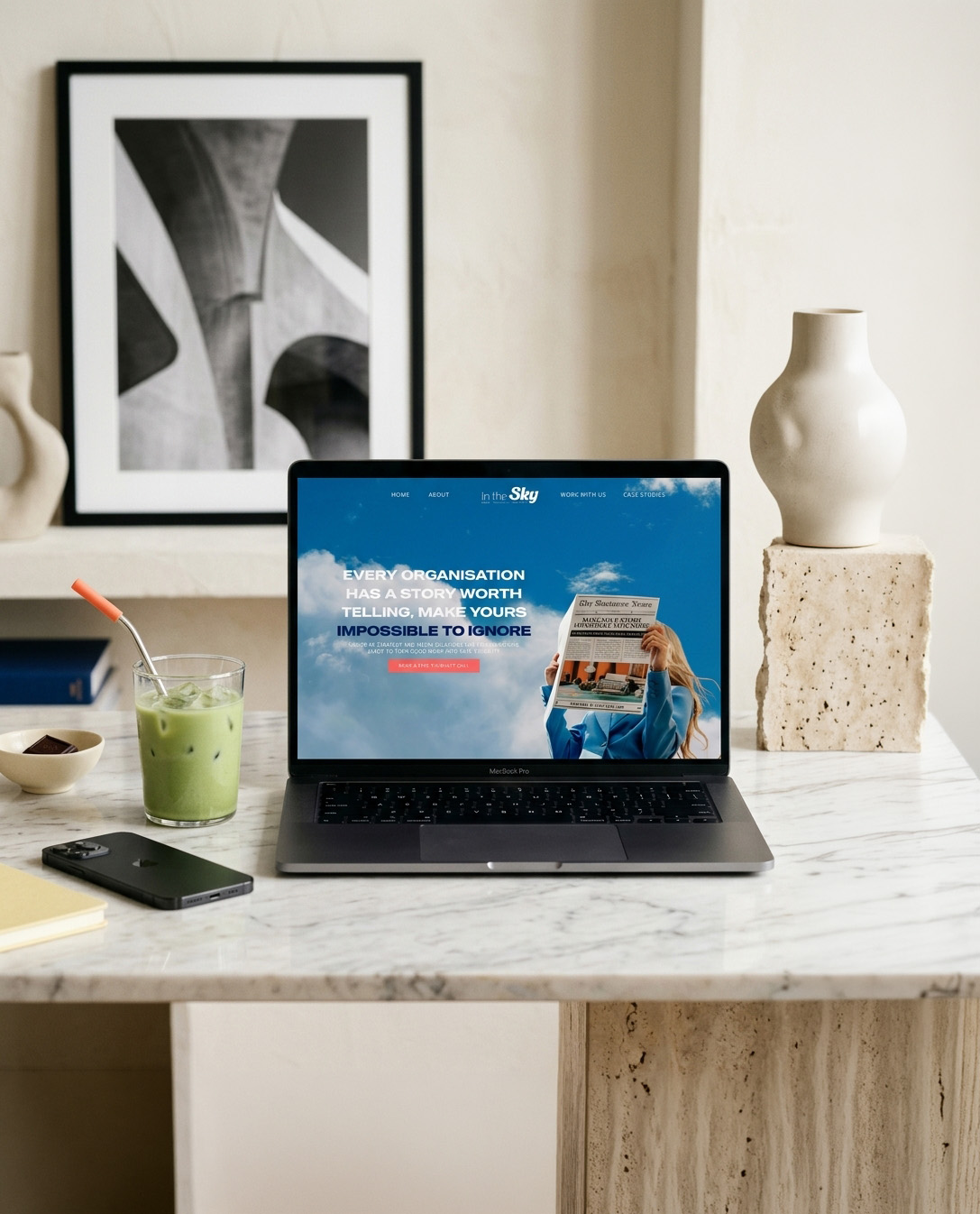 A laptop displaying a Sky marketing webpage sits on a marble table beside a green drink and phone, in a calm, minimalist interior with art and a vase.