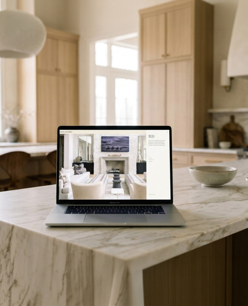 A laptop on a marble kitchen island displays a bright, neutral living room design, surrounded by pale wood cabinetry and soft daylight, creating a calm, refined mood.